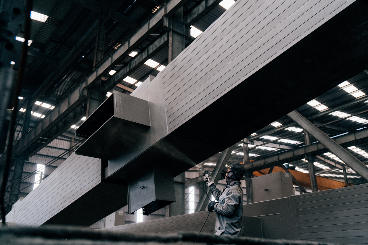 Portfolio A worker sprays paint on large metal beams in an industrial warehouse.