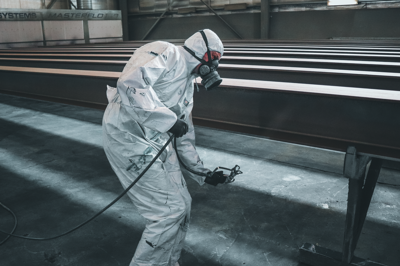 Hakkımızda Worker in protective gear spray painting metal beams inside a factory setting.